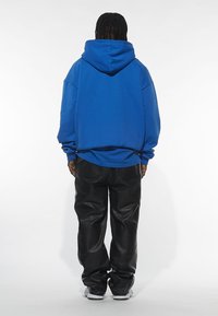Felpa oversize blu con cappuccio, realizzata in tessuto morbido, abbinata a pantaloni in pelle nera. L'insieme presenta una vestibilità comoda e un design minimal.