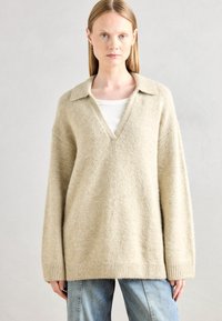 Lys beige oversized sweater i blødt materiale med V-udskæring og krave. Ribbede kanter og lange ærmer, båret over en hvid top.