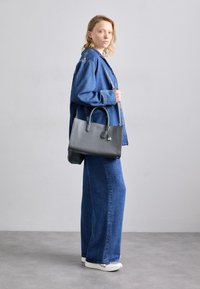 Bolsa de piel gris con superficie texturizada, dos asas y un pequeño detalle de candado. El modelo lleva una chaqueta de mezclilla y jeans de pierna ancha.