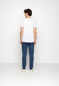 Hvit bomulls T-skjorte med rund hals, kombinert med blå denimjeans. Modellen vender bort, og viser den tettsittende designen og nøytrale farger.