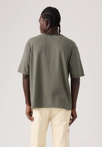 Ριγέ μπλουζάκι με άνετη εφαρμογή σε ελιά και εκρού, με στρογγυλή λαιμόκοψη και κοντά μανίκια, κατασκευασμένο από απαλό βαμβακερό υλικό.