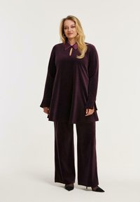 Pluche, diep paarse twee-delige outfit met een lange tuniek met een sleutelgat halslijn en bijpassende wijde broek. Zachte textuur, elegante kraag.