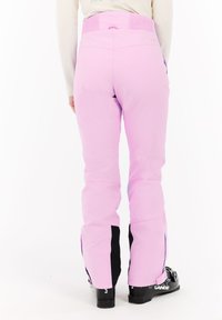 Persoon in roze skibroek en zwarte skischoenen, staand met de rug naar de camera op een witte achtergrond.