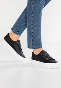 Černé kožené tenisky s kontrastními bílými podrážkami, s texturovanými tkaničkami se stříbrnými akcenty, nošeny s zkrácenými denimovými džínami.