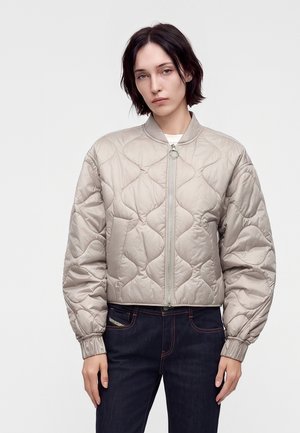 Jeune personne aux cheveux courts et foncés porte une veste bomber matelassée beige clair et un jean foncé sur un fond blanc uni.