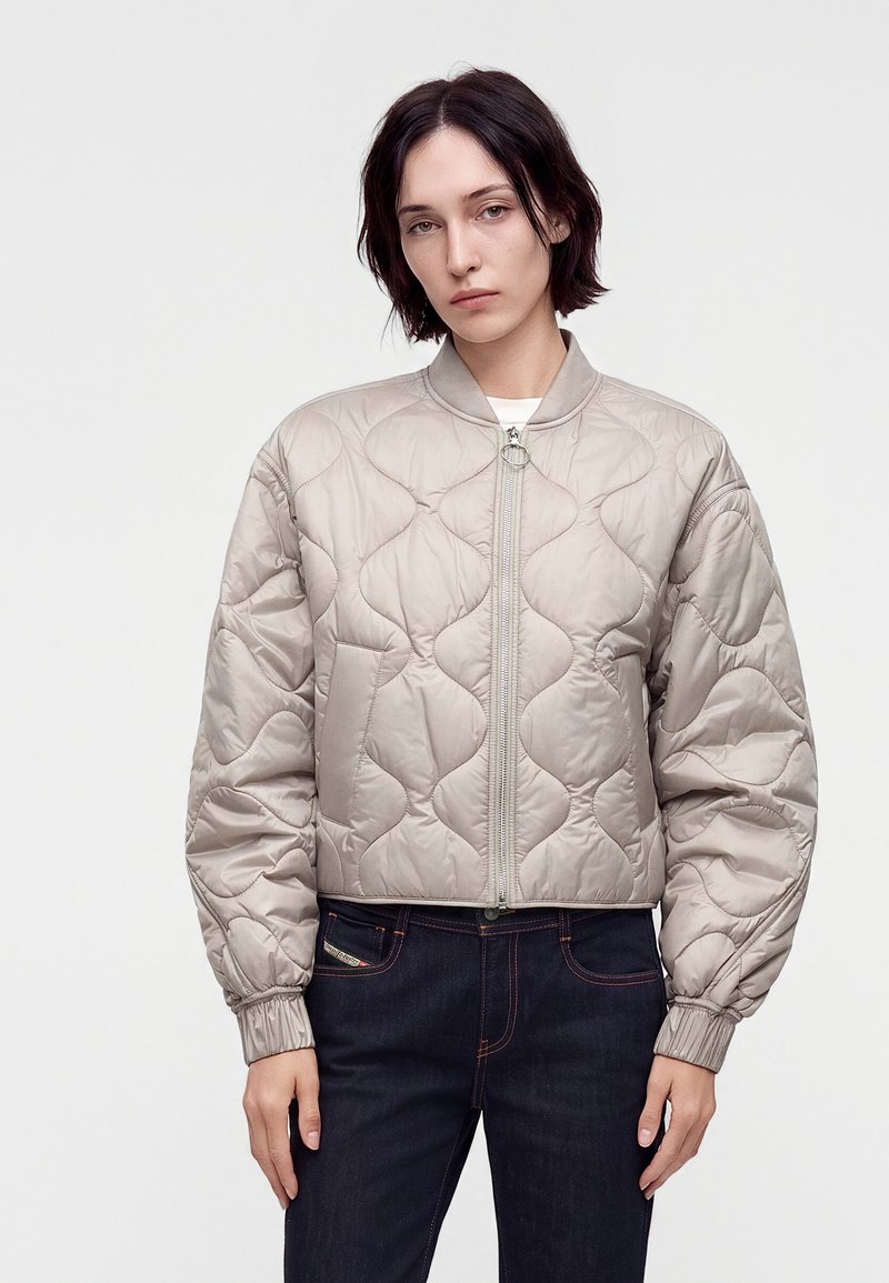 Jeune personne aux cheveux courts et foncés porte une veste bomber matelassée beige clair et un jean foncé sur un fond blanc uni.