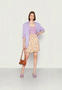 Blazer lilás claro sobre um top lilás, combinado com uma saia amarela com padrão de leopardo, sandálias de salto bloco lilás e uma handbag castanha.