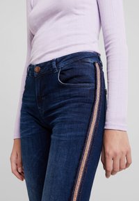 Mörkblå högmidjade jeans med slim fit, med en bronsefärgad sidosträcka och texturerad tyg. Framtida fickor synliga, knäppt med knapp.