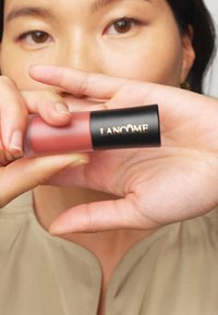 Produkt do ust Lancôme trzymany w widoczny sposób, z matowym różowym kolorem i błyszcząca czarną nakrętką z złotym napisem. Gładka tekstura, kompaktowy design.