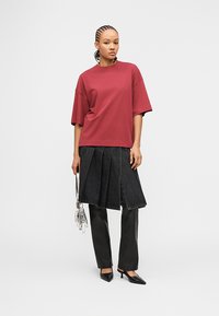 Γυναίκα με πλεξούδες φοράει κόκκινη φαρδιά μπλούζα, μαύρη πλισέ φούστα πάνω από μαύρο παντελόνι, μαύρες γόβες και κρατά μια μικρή ασημένια τσάντα.