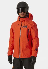 Chaqueta de esquí naranja con cierre frontal, capucha ajustable y dos bolsillos con cremallera. Acentos negros en el logo y en las cremalleras. Acabado mate.