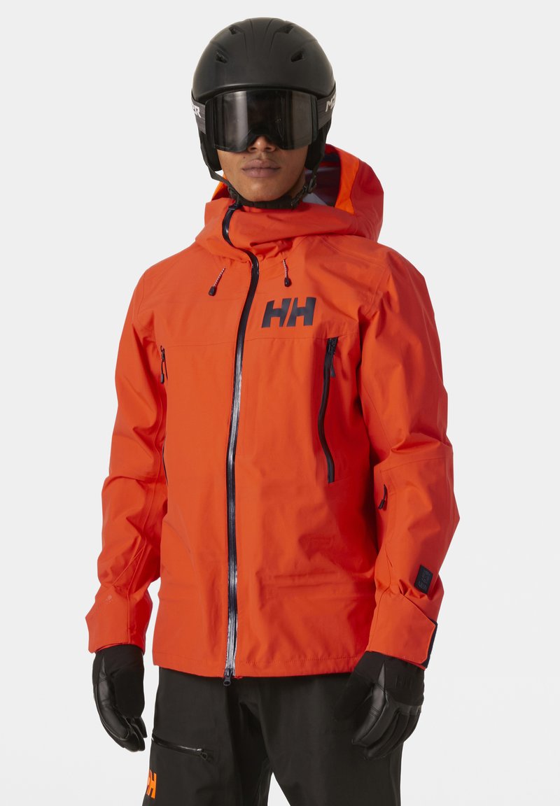 Chaqueta de esquí naranja con cierre frontal, capucha ajustable y dos bolsillos con cremallera. Acentos negros en el logo y en las cremalleras. Acabado mate.