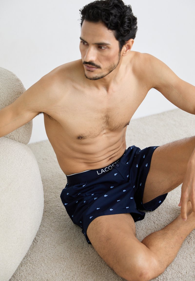 Lacoste navy boxer shorts with light blue logo pattern, elastic waistband, and button closure. Sitting on a textured beige surface.