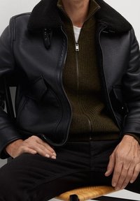 Giacca in pelle nera con colletto in shearling, indossata sopra un maglione verde a zip. Il modello è seduto, con le mani appoggiate sui pantaloni neri.