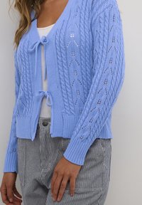 Jasnoniebieski sweter rozpinany na drutach z warkoczowymi wzorami, dekoltem w serek i kokardkami z przodu. Do tego dopasowane, wysokie spodnie w paski.