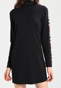 Robe noire à manches longues avec un col montant. Présente un texte en rouge et blanc le long de la manche gauche. Tissu lisse avec une coupe décontractée.