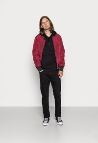Red bomber jacket with black detailing, layered over a black hooded sweatshirt, paired with black pants and black sneakers.
