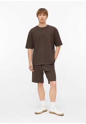 Junger Mann, der ein locker sitzendes dunkelbraunes T-Shirt- und Shorts-Set mit weißen Socken und weißen Sneakers trägt, steht vor einem neutralen Hintergrund.