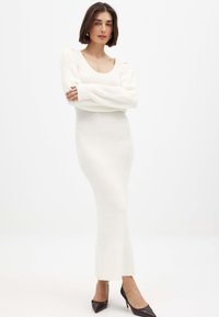 Kootud, pika varrukaga kleit valges, millel on vooderdatud disain, ümar kaelus ja soonikkoes. Koos tumedate kõrgete kontsadega kingadega.