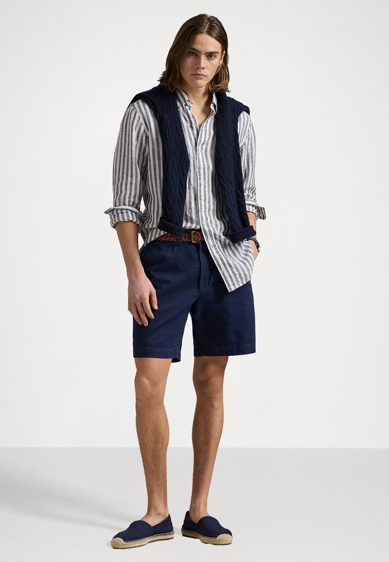 Marineblauwe kabelgebreide mouwloze trui over een gestreept, knoopshirts, gecombineerd met donkere shorts en marineblauwe espadrilles.