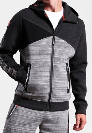 Mężczyzna ubrany w szarą i czarną bluzę rozpinaną na zamek oraz pasujące szare spodnie z logo "Superdry" na rękawach, kieszeniach i spodniach.