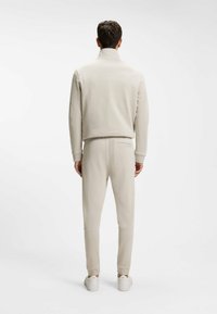 Άνδρας που φοράει μπεζ φούτερ με ψηλό λαιμό και αντίστοιχο παντελόνι φόρμας, στέκεται γυρισμένος με την πλάτη σε μια απλή λευκή φόντο.