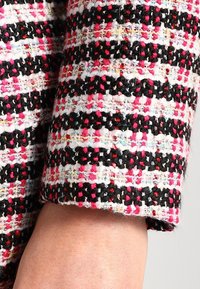 Tissu texturé avec des rayures horizontales en rose, noir, blanc et fils multicolores ; vue rapprochée de la manche et du bras.