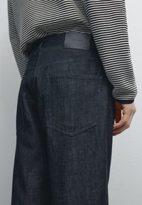 Mörkblå denimjeans med rak benform, med en bakficka och en märkesläderpatch på midjan. Ränder syns på toppen.
