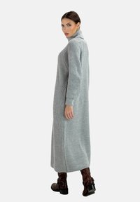 Hellgraues geripptes Strickkleid mit hohem Kragen, langen Ärmeln und seitlichen Schlitzen, kombiniert mit braunen Knöchelstiefeln.