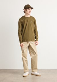 Langärmliges olivgrünes Hemd mit einem quadratischen Logo-Patch, beigen Hosen und weißen Turnschuhen. Das Model trägt eine passende olivgrüne Mütze.
