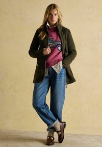 Blondynka w ciemnozielonej kurtce, różowym swetrze z wzorem ciężarówki, zrolowanych niebieskich dżinsach, beżowych skarpetkach i brązowych mokasynach stoi pod beżową ścianą.