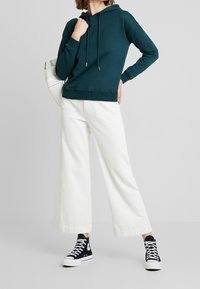 Sudadera con capucha verde azulado y cordones, pantalones de pana anchos blancos y zapatillas altas negras. El conjunto presenta un diseño simple.
