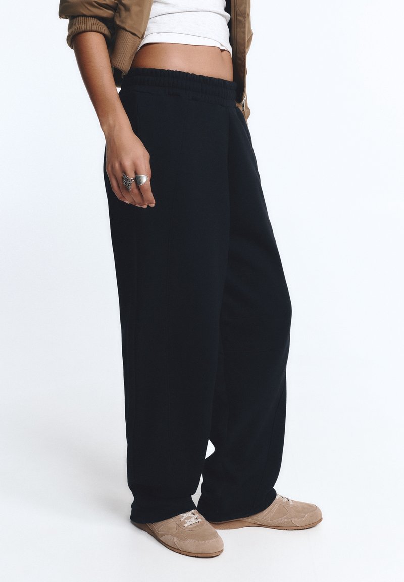 Personne portant un pantalon noir ample, des baskets beiges, un top blanc court et une veste marron, debout devant un fond uni.