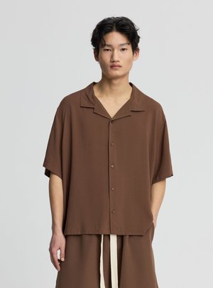 Giovane uomo con capelli corti neri che indossa una camicia marrone ampia con bottoni e pantaloni coordinati con coulisse bianche su sfondo semplice.