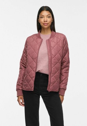 Femme portant une veste matelassée mauve sur un pull rose clair et un pantalon noir, debout devant un fond blanc uni.