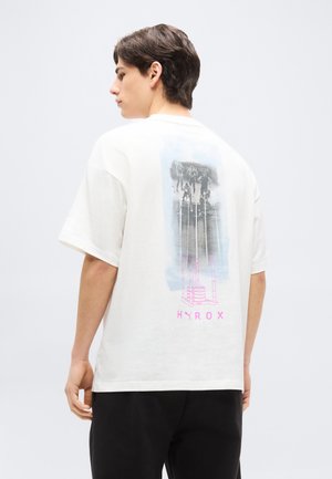 Fiatal férfi fehér pólóban, melyen fekete-fehér futókat ábrázoló grafika és rózsaszín HYROX felirat található a hátoldalon, semleges világos háttér előtt áll.