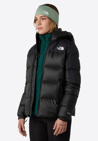 Musta eristetty puffer-takki hupulla ja vetoketjullisilla taskuilla, jossa on kiiltävä pinta ja kontrastivärit hartioilla. Vihreä hiuspanta päässä.