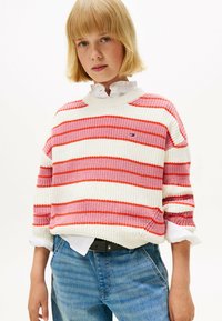 Maglione lavorato a maglia con righe rosse e rosa su sfondo crema; presenta un collo a giro e una vestibilità rilassata, indossato sopra una camicia bianca.