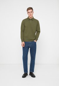 Zielona, długosleeve'owa koszulka polo z trzema guzikami, w połączeniu z niebieskimi dżinsami i czarnymi loafersami. Prosty design, standardowy krój.