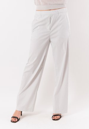 Donna che indossa pantaloni grigio chiaro a vita alta e gamba ampia e sandali neri con tacco e punta aperta, in piedi davanti a uno sfondo bianco semplice.