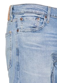 Jean denim bleu clair en gros plan montrant les poches avant et arrière avec des rivets en cuivre et des passants de ceinture sur fond blanc.