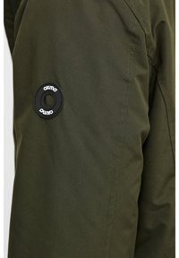 Olivgrön jacka med en texturerad tyg, sid sömstickning och en svart cirkulär logopatch på armen.