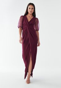 Burgundijas kleita ar grozītu priekšpusi, dziļu v-veida kakla izgriezumu un caurspīdīgām puff piedurknēm, kas rotātas ar maziem punktiem. Izsists apakšdaļa uzlabo kustības.