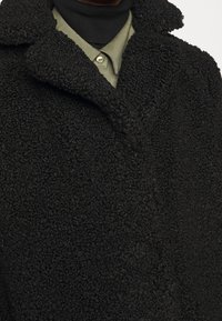 Cappotto in pile nero con texture e ampie revers indossato sopra una camicia verde oliva a bottoni e un dolcevita nero.