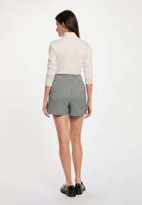 Cremfarbener Langarmoberteil kombiniert mit hochtaillierten Shorts im schwarz-weißen Karomuster. Glatte Textur, figurbetonter Schnitt und flache schwarze Schuhe.