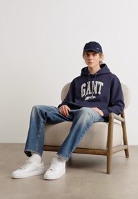 Marinblå hettegenser med hvitt logo, blå jeans og hvite sneakers. Sitter på en lys stoffjakkestol, med en mørk caps. Nøytral bakgrunn.