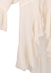 Blusa lunga beige chiaro a maniche lunghe con bordi in pizzo, orli arricciati e nappine. Realizzata in un tessuto leggero e testurizzato.
