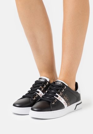 Black leather sneakers with white accents, featuring gold branding and decorative patterns. Lace-up design with a textured sole.