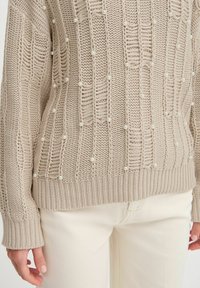 Pull en maille beige avec des rayures texturées et de petites touches de perles blanches. Manches et ourlet côtelés. Porté par-dessus un pantalon de couleur claire.