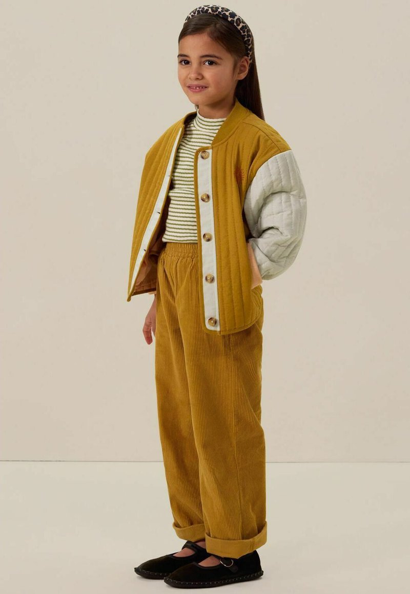 Geel corduroy jasje met witte gestikte mouwen, gestreepte shirt, bijpassende gele corduroy broek en zwarte instappers.